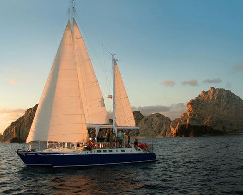 A sailing catamaran under sail at sunset near the Land's End rocks in Cabo San Lucas